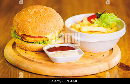 Burger with cheese meat and salad. Cheat meal. Delicious burger with sesame seeds. Burger menu. High calorie snack. Hamburger and french fries and tomato sauce on wooden board. Fast food concept. Stock Photo