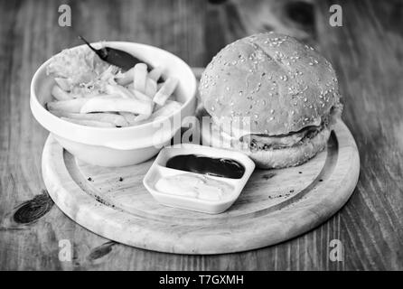 Delicious burger with sesame seeds. Burger menu. High calorie snack. Hamburger and french fries and tomato sauce on wooden board. Fast food concept. Burger with cheese meat and salad. Cheat meal. Stock Photo