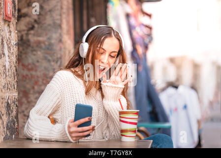 Beautiful attractive young trendy girl with headphones,smartphone and sunglasses talks on phone and waves hand while sitting a street cafe Stock Photo