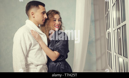 Portrait of happy young couple in love in bathrobes are looking out the window and hugging, toned Stock Photo