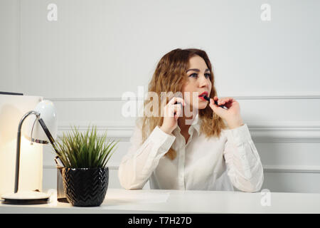 Young curly-haired woman secretary is talking on mobile phone with client and taking notes sitting at table in the office. Office working concept. Stock Photo