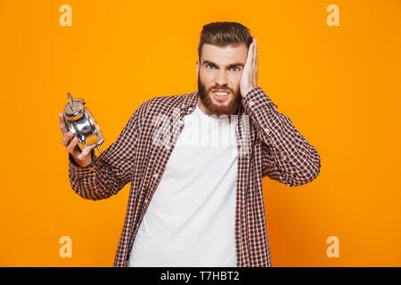 Portrait of a shocked young man wearing casual clothes standing isolated over yellow background, holding alarm clock Stock Photo