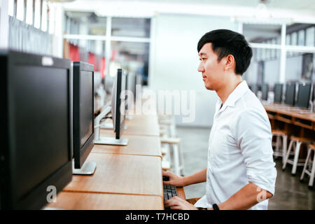 Side view of casual Asian man using computer at desk in modern classroom Stock Photo