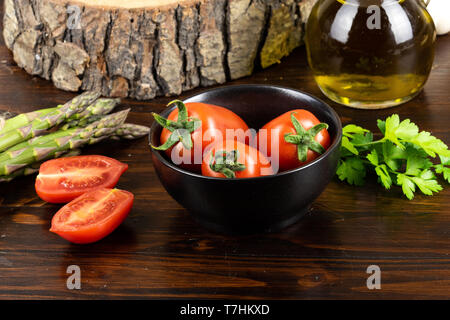 tomatoes in a black bowl on a dark wooden table and asparagus and parsley Stock Photo