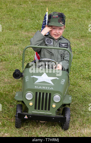 A young boy dressed in an overall of the 82nd Airborne Division saluting whilst driving a Willys jeep at the D-Day celebrations in Normandy, France Stock Photo