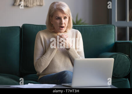 Shocked grey haired mature woman reading unexpected news on laptop Stock Photo