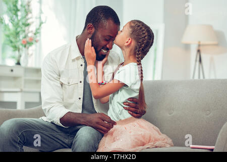 Little daughter lovingly kissing her Afro-American handsome dad forehead. Stock Photo