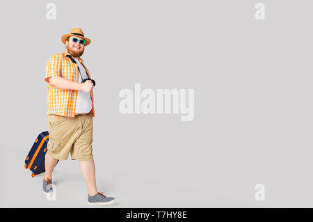 Fat funny man with a suitcase smiling on a gray background. Stock Photo