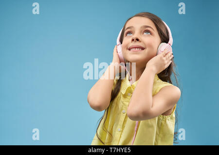 Beautiful, young girl with long hair holding hands pink headphones and listening music. Happy, cute child in yellow blouse looking up and posing in studio on blue background. Stock Photo