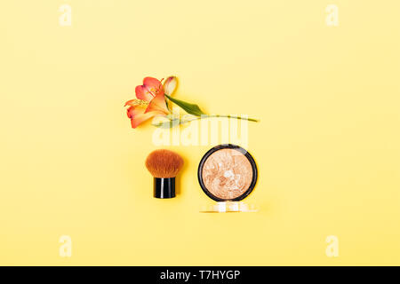 Compact bronzer powder and kabuki brush next to flower on yellow background, top view. Beauty product and tool for summer glow makeup, flat lay. Stock Photo