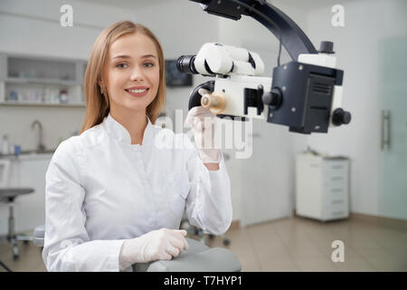 Happy female dentist posing in dentistry private clinic. Beautiful and charming blonde woman looking at camera and smiling, Stomatologist holding hand on dental microscope. Stock Photo