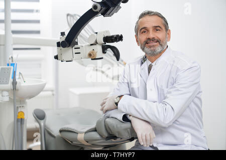Mature stomatologist posing on workplace, near dental microscope, leaning on dentist chair. Handsome, bearded stomatologist wearing in white medical coat. Man looking at camera and smiling. Stock Photo