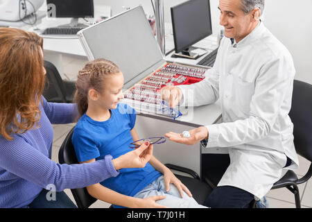 Smiling doctor ophthalmologist sitting at table with eye test lens, holding and offering contact lenses or glasses for patient. Pretty girl looking at container, choosing. Stock Photo