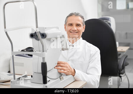 Doctor optometrist wearing in white medical coat working on special ophthalmology equipment in optician office. Correction and treatment of vision with modern slit lamp machine. Stock Photo