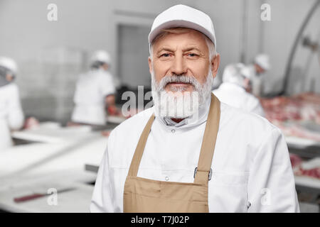 Elderly man working on sausage production, factory. Professional in white uniform, white cap and brown apron standing, posing and looking at camera. Meat manufacturing. Stock Photo
