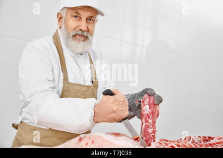 Handsome elderly man working as butcher holding knife and meat, looking at camera. Worker wearing in white uniform, brown apron and special gloves cutting fresh pork carcasses. Stock Photo