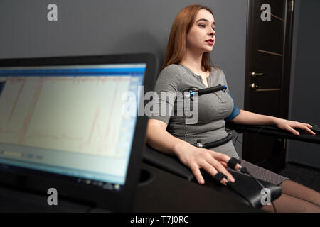 Young beautiful woman sitting with attached sensors on fingers, hands and body, testing with computer polygraph, looking away. Graph and polygraph  on screen of lie detector. Stock Photo