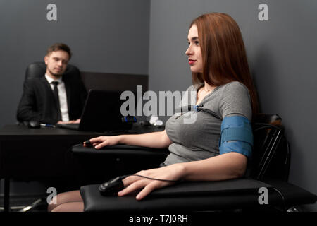 Beautiful, serious woman sitting in grey room and examinating on lie detector. Finger sensors, electrodes, wire on hands and body. Young man testing woman with computer polygraph. Stock Photo