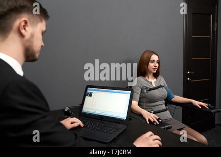 Concentrated woman sitting in grey room, testing at computer polygraph. Young man sitting at table and looking screen of polygraph and monitoring polygraph . Stock Photo