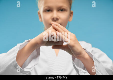 Selective focus of little hands of girl showing fist in studio. Cute female child looking at camera and posing on blue isolated background. Young fighter training and doing karate. Concept of sport. Stock Photo