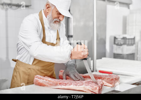 Elderly man working on meat production. Professional butcher in white uniform, cap, brown apron and special gloves standing near table, holding knife and cutting meat. Food industry. Stock Photo