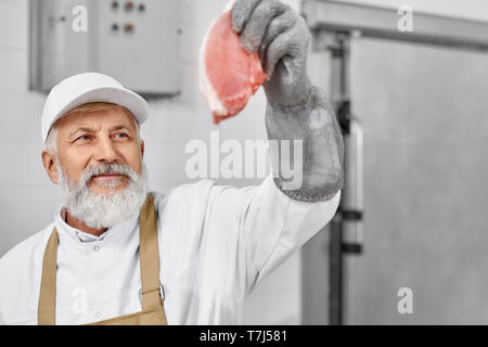 Butcher wearing in white uniform, white cap, brown apron, special gloves holding steak, checking quality. Elderly man working on meat production, modern factory. Food industry. Stock Photo
