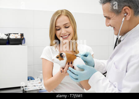 Happy woman looking at her pet, smiling. Owner holding hamster and veterinarian observing animal with stethoscope. Small hamster on examination in professional vet clinic. Stock Photo