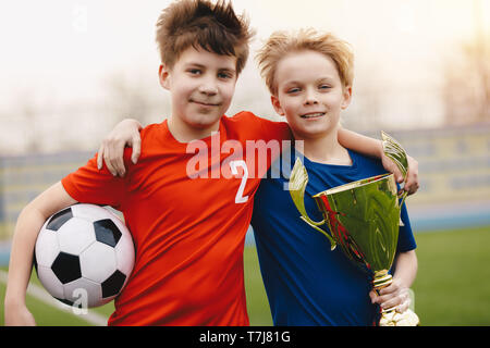 Two happy boys soccer players holding soccer ball and golden trophy. Children football players on the field in red and blue jersey sportswear Stock Photo