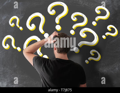 rear view of puzzled man in t-shirt scratching his head in confusion against blackboard filled with question marks Stock Photo