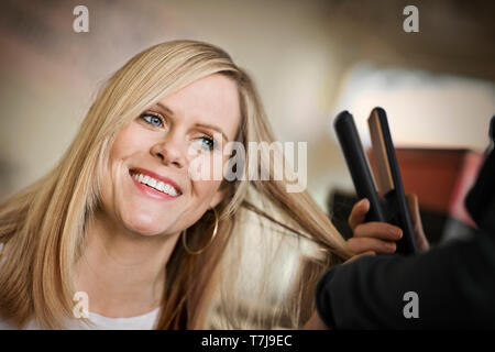 Smiling young woman getting her long hair straightened with a hair iron. Stock Photo