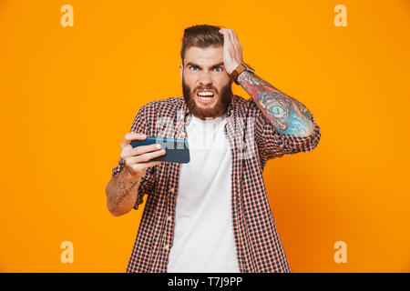 Portrait of an angry young man wearing casual clothes standing isolated over yellow background, playing games on mobile phone Stock Photo