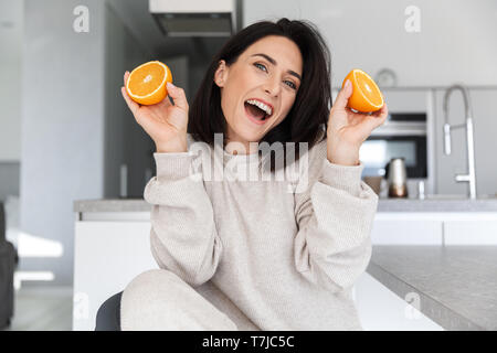 Image of joyful woman 30s holding two pieces of orange while sitting in living room Stock Photo