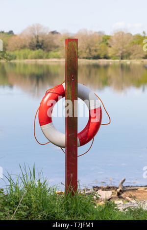 A red and white life saver ring hangs on a red post on the shore of a reservoir. Orange rope hangs loosely around the ring. Stock Photo