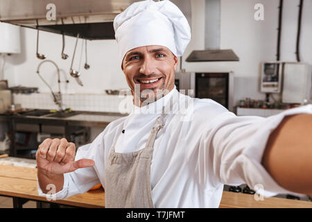 Photo of smiling male chief in apron taking selfie while standing at kitchen in restaurant Stock Photo