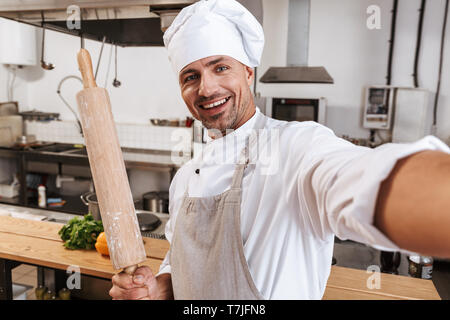 Photo of handsome man chief in apron taking selfie while standing at kitchen in restaurant Stock Photo