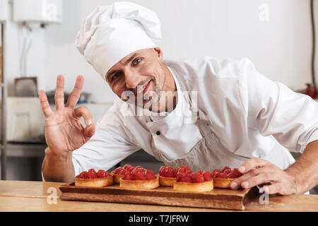 Photo of smiling male chief in white uniform holding plate with cakes while cooking at kitchen in restaurant Stock Photo