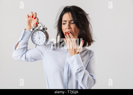 Tired businesswoman smartly dressed standing isolated over white background, showing alarm clock, yawning Stock Photo