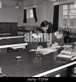 1960s, historical, in an empty secondary school classroom, a female mathematics teacher overseeing the study or book work of two male pupils, who are doing extra study or revision after class, South London, England, UK. Stock Photo