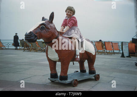 1960, historical, happy little girl sitting on a large seaside wheeled toy horse beside the coast at Brighton, W. Sussex, England, UK. Stock Photo