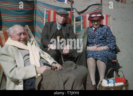 1960s, historical, three elderly people in their normal everyday clothing sitting together outdoors on striped deckchairs on a sandy beach, England, UK. Beside the lady is a basket with a picnic inside. Stock Photo