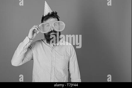 Man with beard and mustache on calm face lonely on his birthday, blue background. Guy in party hat celebrates alone. Birthday concept. Hipster in giant sunglasses celebrating birthday, copy space. Stock Photo