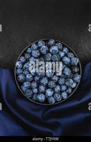 Blueberries in round and black cast iron bowl on blue fabric and black stone background with copy space Stock Photo