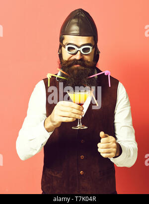 handsome bearded pilot or aviator man with long beard and mustache on serious face holding glass of alcoholic beverage in vintage suede leather waistcoat with hat and glasses on red studio background. Stock Photo