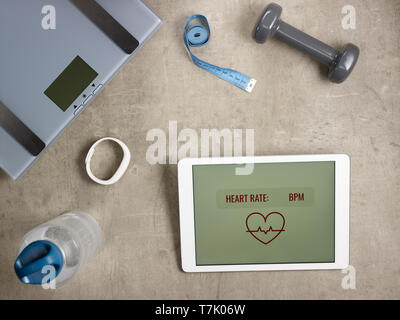Closeup on weight scales, grey dumbbell, white fitness tracker, bottle of water, tape measure and tablet PC with heart rate app laying on the floor. Stock Photo