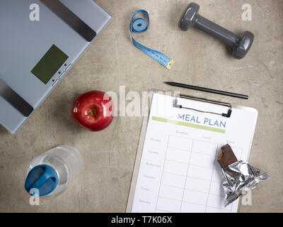 Closeup on weight scales, grey dumbbell, red apple, bottle of water, tape measure, bitten raw protein bar, black pen and clipboard with meal plan layi Stock Photo