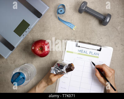 Closeup on weight scales, grey dumbbell, red apple, bottle of water, tape measure laying on the floor and female hands with bitten raw protein bar fil Stock Photo