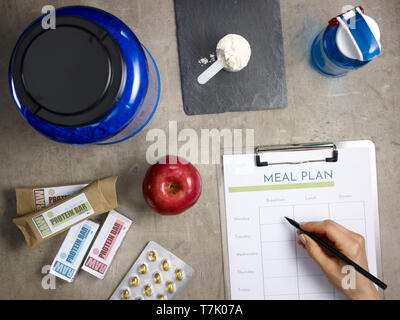 Closeup on big blue protein jar, shaker, measuring spoon with powder, raw protein bars, red apple, sport supplements in tablets and female hand with b Stock Photo