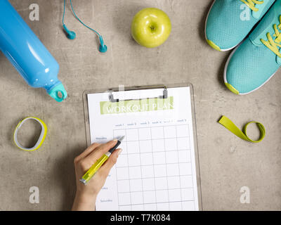 Closeup on green apple, headphones, elastic band, sneakers, fitness tracker, bottle of water laying on the floor and female hand filling workout plan  Stock Photo