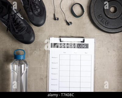 Closeup on laying on the floor black sneakers, headphones, fitness tracker, bottle of water, weight plate and workout log in clipboard. Stock Photo