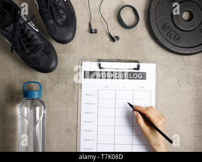 Closeup on laying on the floor black sneakers, headphones, fitness tracker, bottle of water, weight plate and female hand filling workout log in clipb Stock Photo